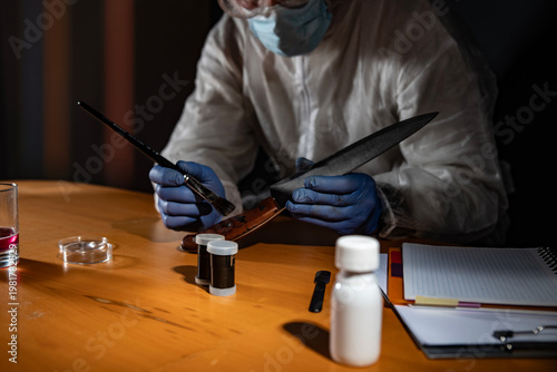 Foto Crime scene evidence processing on wooden desk: gloved hands use a brush to reveal latent prints on a drinking glass beside a knife with blood stains and sample vials