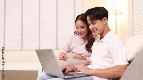 Happy young Asian couple smiles while sitting on a cozy white sofa, looking at a laptop screen together in their living room, perfect for home lifestyle, remote work, or online shopping concepts