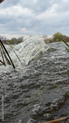 Close-up of turbulent river waves crashing against the shore, splashing through reeds and grass under a cloudy sky. Dynamic water motion with bubbles 