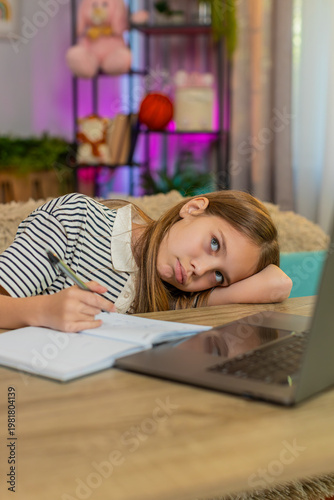 Young child girl sitting at table on home sofa, feeling tired lazy, doing homework with laptop and notebook. School kid slowly writes in exercise book struggling to stay focused during online learning
