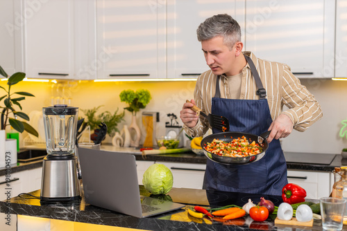 Middle-aged man fries vegetables in pan while watching laptop for diet meal prep in modern kitchen. Young adult guy focuses on healthy food routine during home cooking with serious focused face daily.