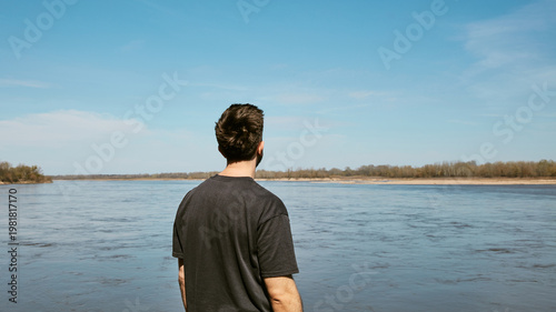 Rear View of Man Looking at Wide River Under Blue Sky