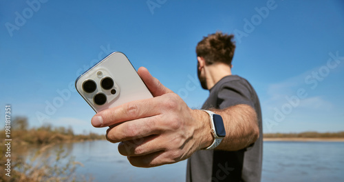 Close up of a man holding a modern white smartphone with three cameras while standing outdoors near a river