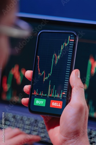 Obraz na plátně Over the shoulder view of a trader closely examining cryptocurrency charts on a smartphone via a trading app, with stock market data displayed on a laptop in the background