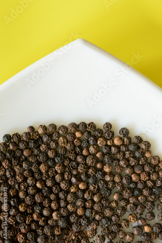 VISTA PARCIAL DE SEMILLAS DE PIMIENTA NEGRA (Piper Nigrum) EN UN PLATO BLANCO SOBRE FONDO AMARILLO