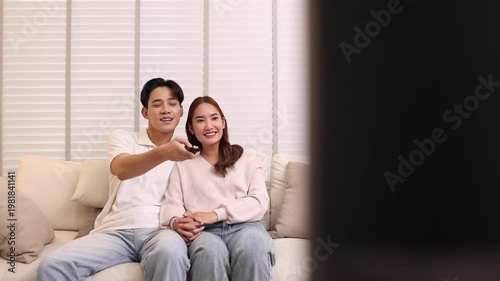 A cheerful young Asian couple relaxes on a sofa, happily pointing a remote control at a television in their cozy living room, perfect for home entertainment, relaxation, or television advertising conc