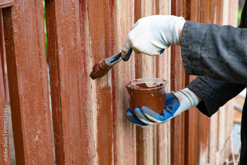 Person painting wooden fence with brown paint in a garden on a sunny day using a brush and a container