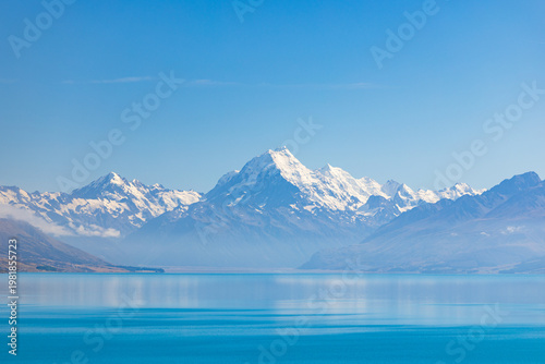View of snow-capped mountains mirrored on the tranquil, turquoise lake, under a clear blue sky, a serene landscape evoking peace and adventure, Lake Pukaki, Mount Cook Lookout, New Zealand.