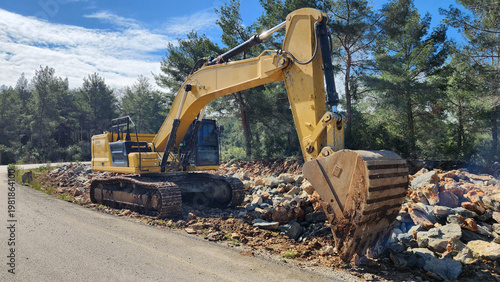 A large yellow excavator is stationary, with its bucket lowered toward a pile of loose rocks and dirt on a steep embankment next to the road