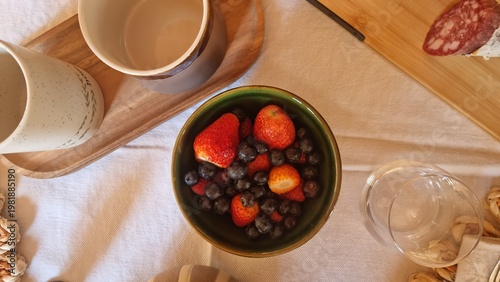 Breakfast setup with bowl of berries, coffee cups, pastries, and sliced salami on wooden table.