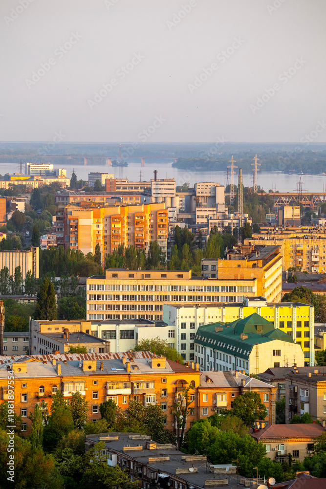 Fototapeta premium Dnipro, Ukraine, cityscape at golden hour with the Dnieper River and residential buildings, industrial structures, and a bridge under a hazy sky, capturing urban life and nature blend Includes copy sp