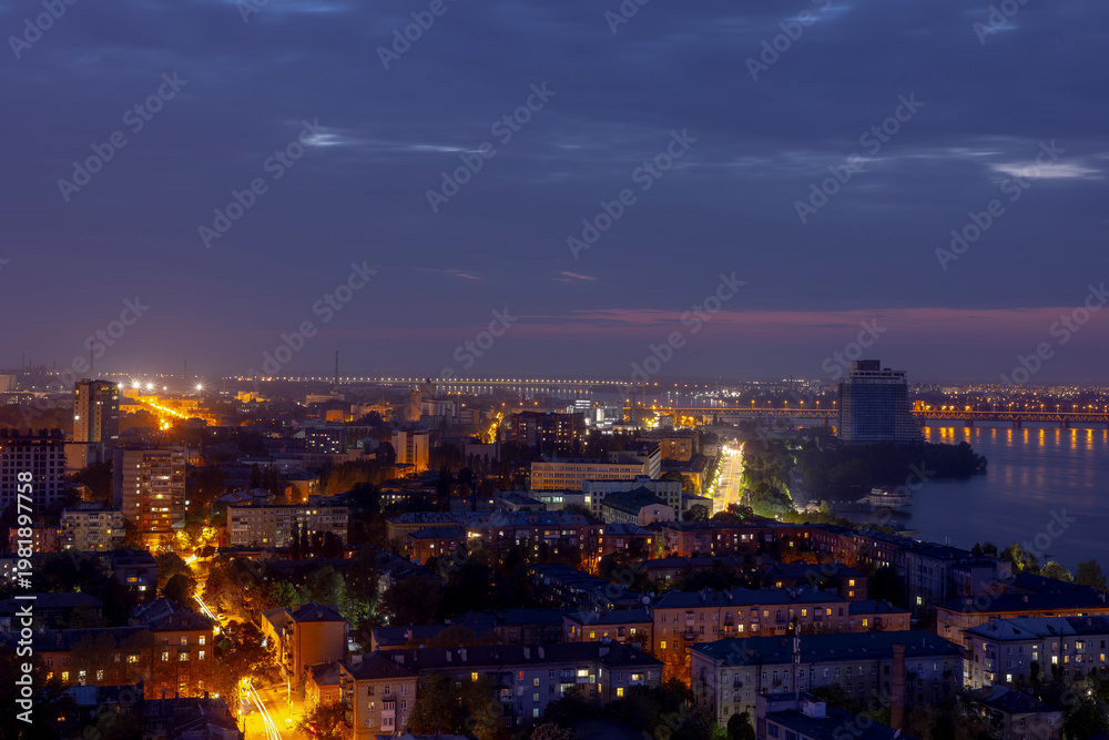 Obraz premium Dnipro, Ukraine, vibrant cityscape at dusk with illuminated bridges spanning the river, showcasing a mix of modern and historical architecture, bustling streets with light trails, and a distant indust