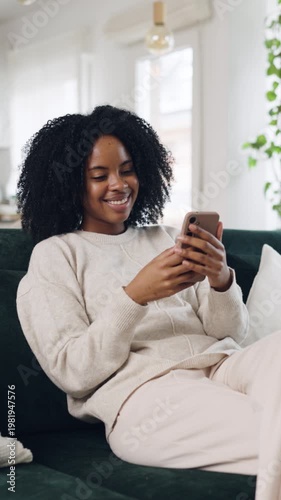 Beautiful african american woman relaxing on a cozy sofa, happily typing and scrolling on her smartphone while enjoying her leisure time at home. Vertical