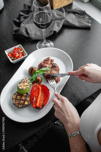 Woman cutting grilled steak served with roasted pepper, zucchini, eggplant, mushrooms, basil, tomato salsa and a glass of red wine