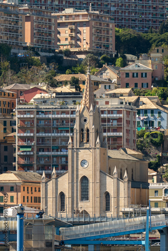 A neo-Gothic church with a slender spire stands prominently in the foreground, contrasted against the densely packed residential apartment blocks climbing the hills of Genoa.
