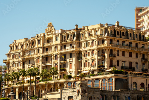 A grand, ornate Mediterranean hotel building stands under a clear sky, featuring classic balconies, intricate carvings, and a row of palm trees along its base.

