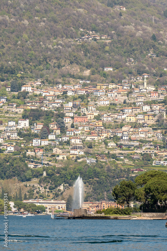 A tall water fountain erupts from a lake in front of a densely populated hillside town.

