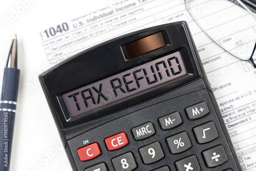 A black calculator displaying TAX REFUND sits atop a 1040 U.S. Individual Income Tax Return form, accompanied by a blue pen and eyeglasses.