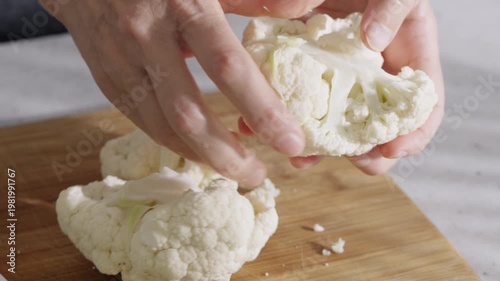 Close up of hands cutting cauliflower into small florets on a wooden board during kitchen food preparation.