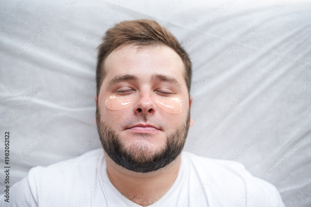 Fototapeta premium Man with under-eye patches during skincare treatment. Close-up of male face with hydrogel eye masks for hydration, anti-aging and dark circles reduction in clinic.
