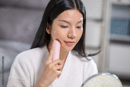 Obraz na płótnie Calm young Asian woman in white bathrobe using facial massager device on cheek l
