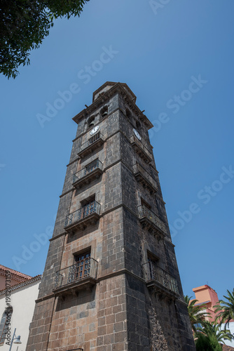 The bell tower of the Church of Nuestra Señora de la Concepción rises against a clear blue sky in La Laguna, Tenerife, Spain, showcasing its historic stone architecture and clock faces