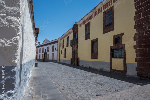 Narrow cobblestone street in La Laguna, Tenerife, lined with historic buildings featuring colorful facades, wooden windows, and traditional Canarian architectural details under a clear blue sky