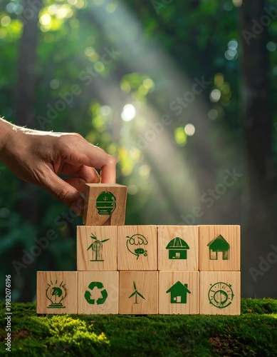 A hand carefully places a wooden block with a leaf icon atop a tower of eco-friendly symbols, representing sustainable development and environmental responsibility within a lush forest setting.