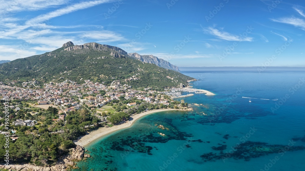 Obraz premium Aerial drone view of Santa Maria Navarrese with marina harbor, turquoise waters, sandy beach and dramatic limestone mountains along the eastern Sardinian coastline on a sunny day.