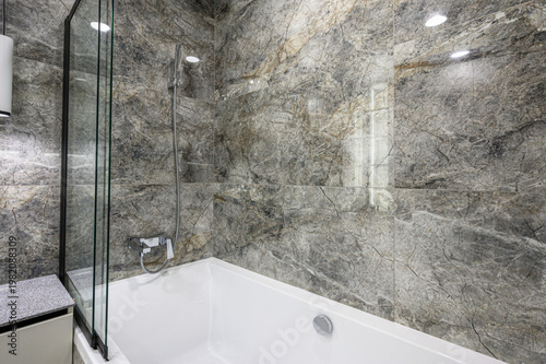 Fotografie modern bathroom with a white bathtub, glass shower screen, grey marble-patterned walls, chrome fixtures and recessed lighting