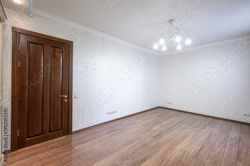 spacious, empty room with light patterned wallpaper, warm wooden laminate flooring, a dark wooden door, and a white chandelier with six lights
