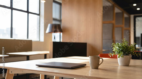 Modern office interior with a closed laptop, coffee mug, and plant on a light wooden desk near a window.