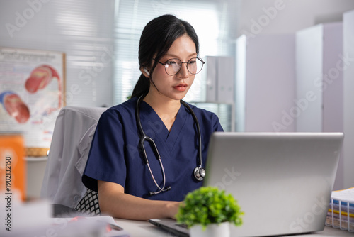 Focused young asian female doctor wearing blue scrub and stethoscope working attentively on laptop computer in bright hospital clinic office reviewing medical patient record data online