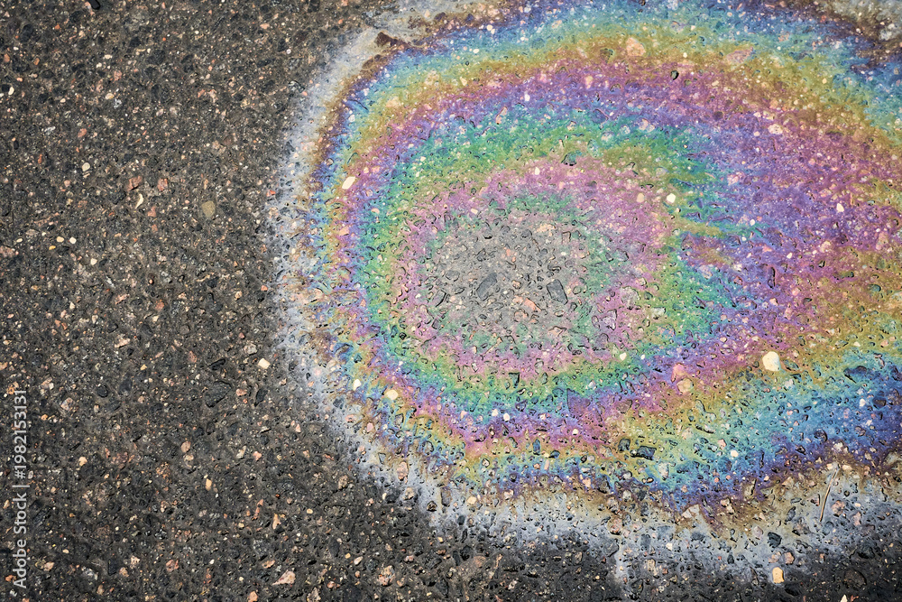 Obraz premium Iridescent oil slick creating colorful rainbow pattern on dark asphalt, symbolizing fuel pollution and liquid waste