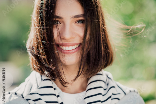 Smiling young woman portrait outdoors in natural light with short brown hair and striped sweater, happy casual lifestyle close-up showing bright smile and fresh spring atmosphere