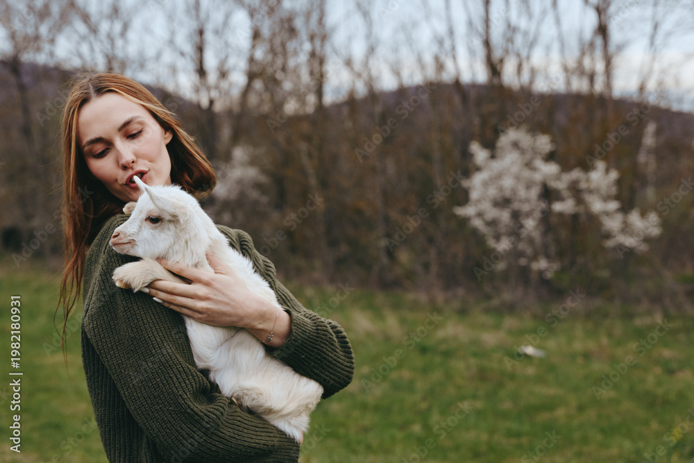 Obraz premium Caring woman holds a white goat kid outdoors in a park, sharing a tender moment. Lifestyle scene emphasizes nature, gentleness, and bonding, with soft natural light creating a calm, warm mood.