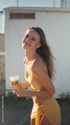 Beautiful happy woman dancing on the beach and drinking a fresh mojito cocktail during sunset