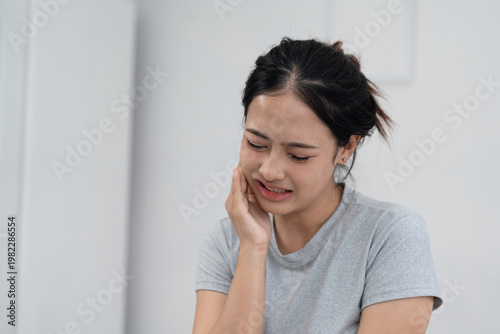 Young Asian woman suffering from toothache, touching cheek with pain, dental problem, sensitivity or cavity, oral health issue and healthcare concept in home environment