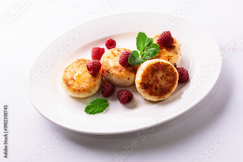 Cottage cheese pancakes with honey, raspberry and mint leaves on white ceramic plate on white marble table top view. Healthy homemade dessert.