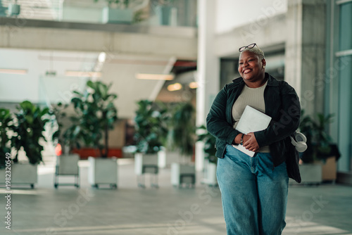 Quadro su tela Black businesswoman smiling, carrying laptop in modern office, portraying succes