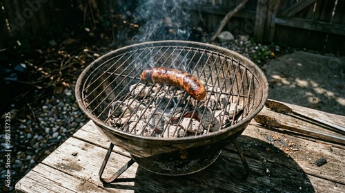 Grilled sausage on a charcoal barbecue grill with smoke rising, placed on a wooden table outdoors in a backyard setting surrounded by greenery
