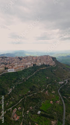 Aerial view of Enna, Sicily, showcasing the historic town perched on a hill surrounded by lush green valleys and winding roads under a cloudy sky. Vertical