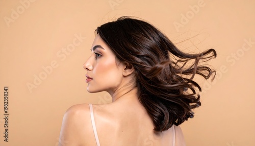 Brunette woman with loose waves looks left over her shoulder against a tan backdrop, revealing bare skin