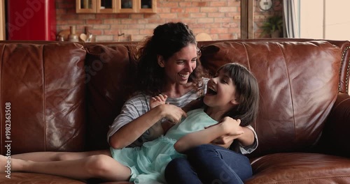 Loving mother playing with child daughter on couch, tickling, laughing, enjoying playtime together on leisure time in living room. Family having fun at home, wellbeing, motherhood, carefree childhood