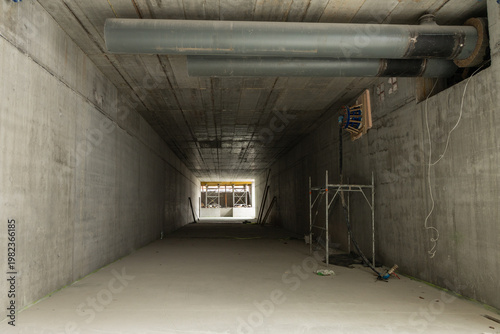Wide-angle view inside a raw, unfinished concrete pedestrian underpass.. Raw concrete surfaces, exposed horizontal pipes with valves and a gauge panel, scaffolding, and a large opening with strong