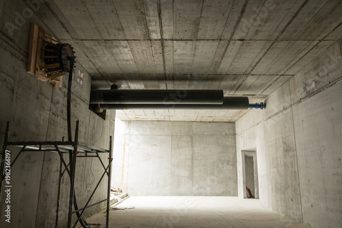 Wide-angle view inside a raw, unfinished concrete pedestrian underpass.. Raw concrete surfaces, exposed horizontal pipes with valves and a gauge panel, scaffolding, and a large opening with strong