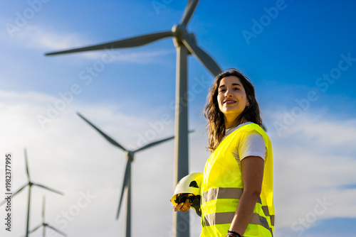 Woman engineer holding helmet and smiling, standing in front of wind turbines