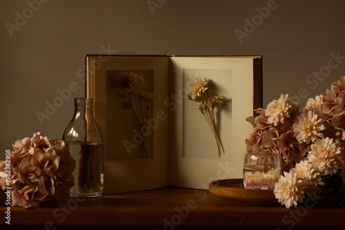 Photos A serene still life with an antique book displaying pressed chrysanthemums, surrounded by elegant silk flowers and a glass bottle