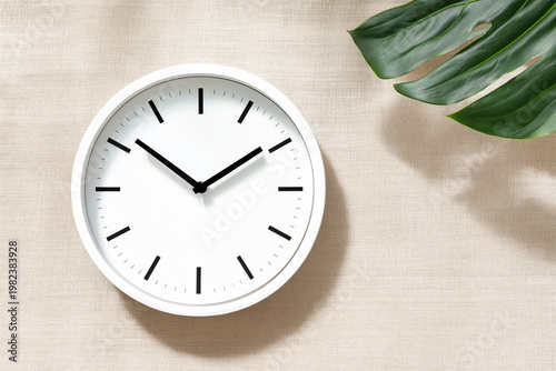 Minimalist white clock with monstera leaf on beige linen texture background for home decor concept.