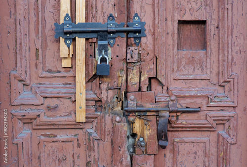 Very old damaged wooden entrance door with peeling paint, broken panels, temporary repairs, new padlock and rusty latches, front view of insecure neglected property access detail, no people